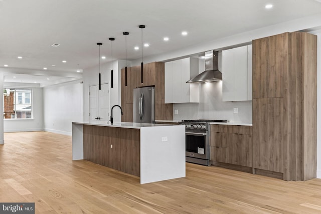 kitchen with wall chimney range hood, a center island with sink, light wood-type flooring, appliances with stainless steel finishes, and modern cabinets