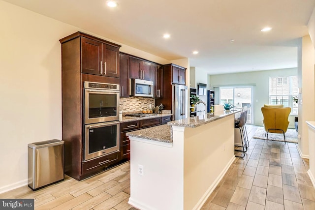 kitchen with a breakfast bar area, stainless steel appliances, light stone countertops, a kitchen island with sink, and decorative backsplash