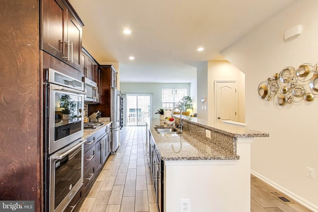 kitchen with sink, dark brown cabinets, stainless steel appliances, light stone countertops, and a kitchen island with sink