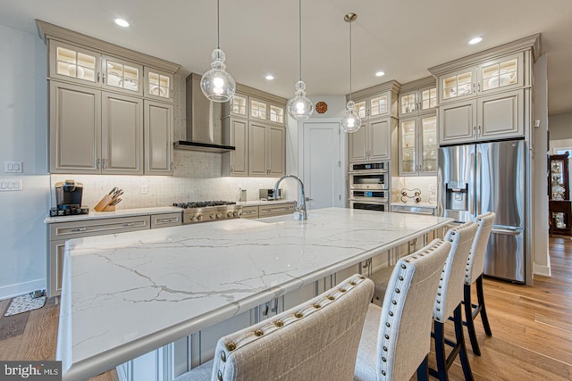 kitchen featuring wall chimney range hood, sink, hanging light fixtures, stainless steel appliances, and a center island with sink