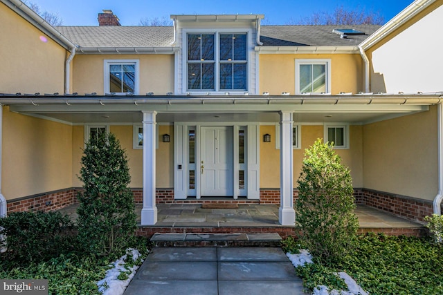 doorway to property with covered porch, a chimney, brick siding, and stucco siding