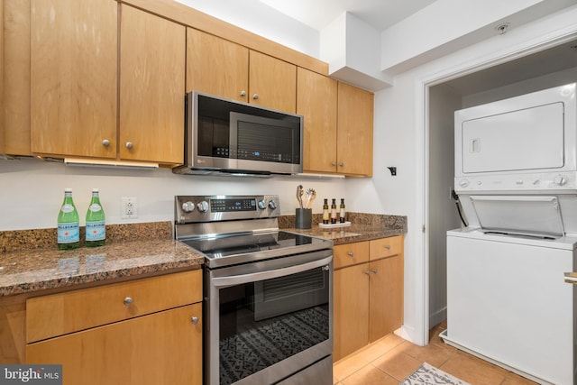 kitchen with stainless steel appliances, stacked washing maching and dryer, light tile patterned floors, and dark stone countertops