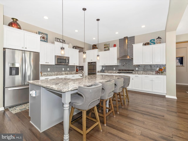 kitchen with dark wood-type flooring, stainless steel appliances, a large island with sink, wall chimney range hood, and white cabinetry