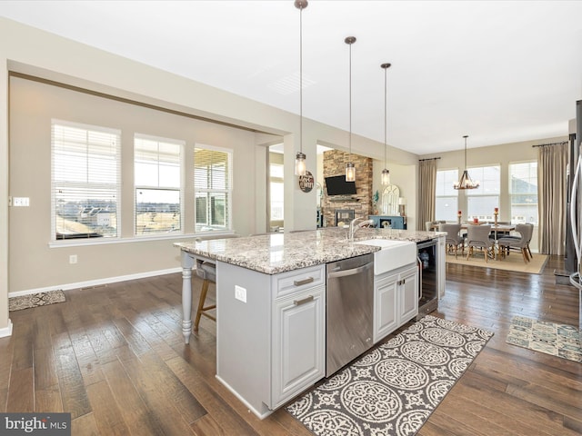 kitchen featuring stainless steel dishwasher, dark wood-type flooring, a sink, a stone fireplace, and an island with sink