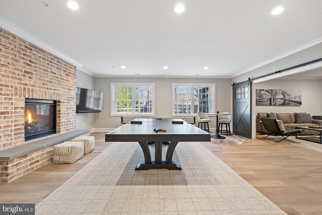 rec room with crown molding, a barn door, a brick fireplace, and light hardwood / wood-style flooring