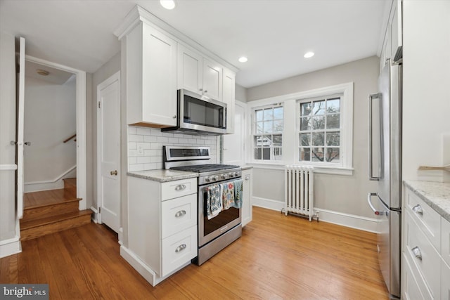 kitchen with appliances with stainless steel finishes, radiator heating unit, white cabinetry, light hardwood / wood-style floors, and light stone countertops