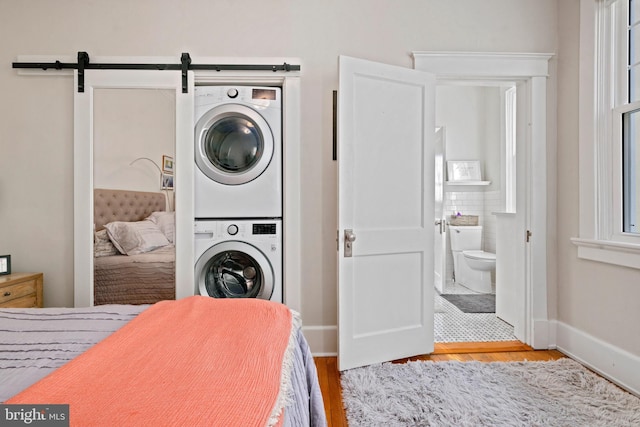 bedroom featuring ensuite bathroom, a barn door, light wood-style flooring, baseboards, and stacked washer and clothes dryer