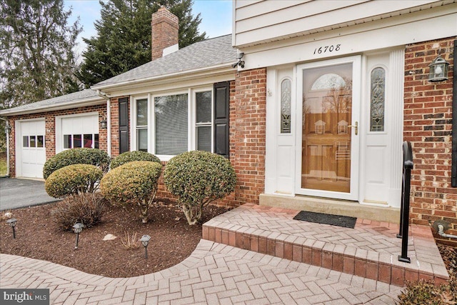 property entrance featuring aphalt driveway, brick siding, a chimney, a shingled roof, and an attached garage