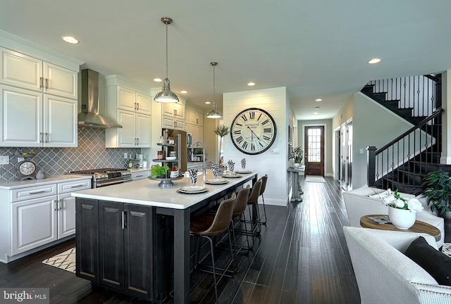 kitchen featuring wall chimney exhaust hood, decorative light fixtures, stainless steel range, an island with sink, and white cabinets