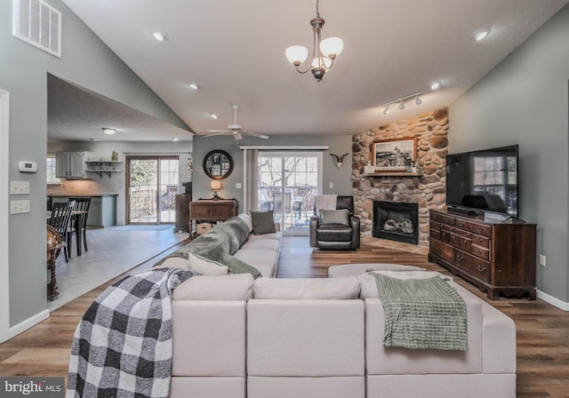 living room featuring vaulted ceiling, a stone fireplace, wood-type flooring, and ceiling fan with notable chandelier