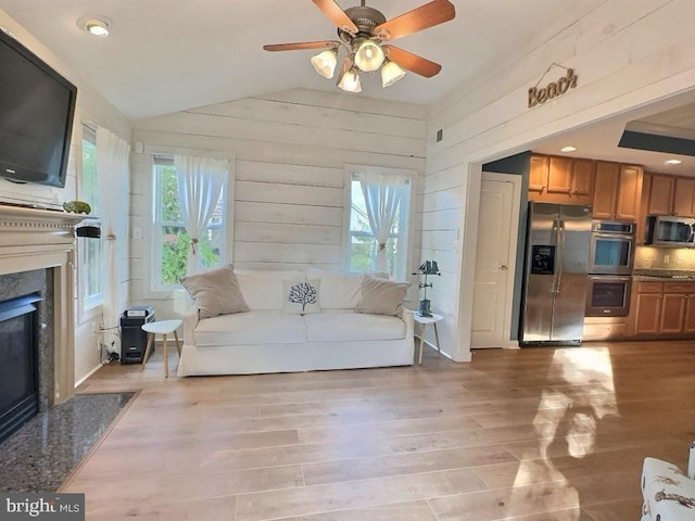 living room with wooden walls, lofted ceiling, ceiling fan, a premium fireplace, and light hardwood / wood-style flooring