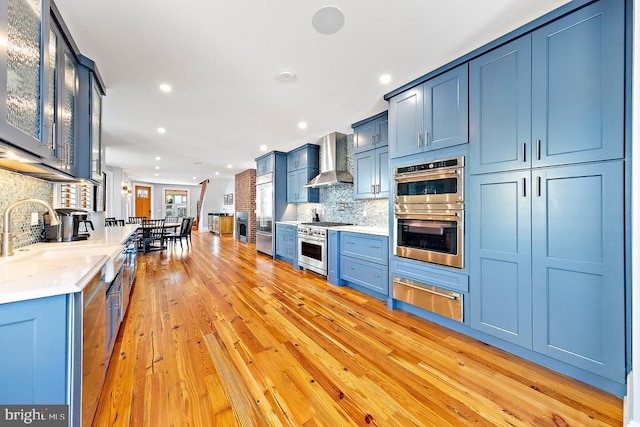 kitchen featuring appliances with stainless steel finishes, wall chimney exhaust hood, blue cabinets, and light hardwood / wood-style floors