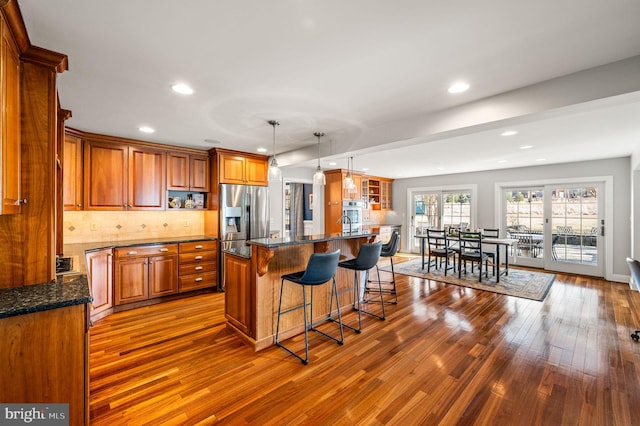 kitchen with tasteful backsplash, hanging light fixtures, brown cabinetry, an island with sink, and stainless steel fridge with ice dispenser