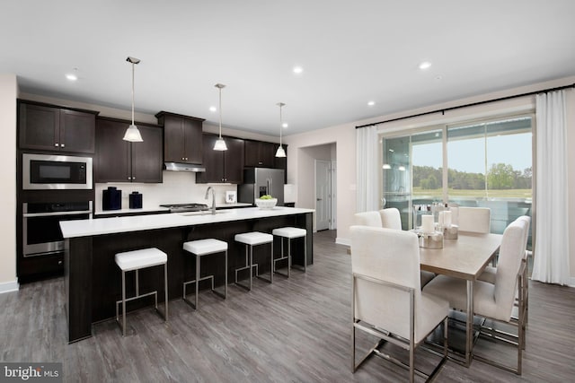 kitchen featuring a center island with sink, light countertops, dark brown cabinetry, under cabinet range hood, and appliances with stainless steel finishes