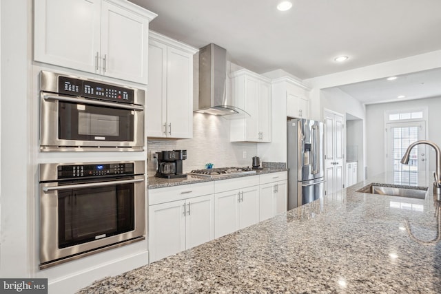 kitchen with backsplash, stainless steel appliances, white cabinetry, wall chimney exhaust hood, and a sink