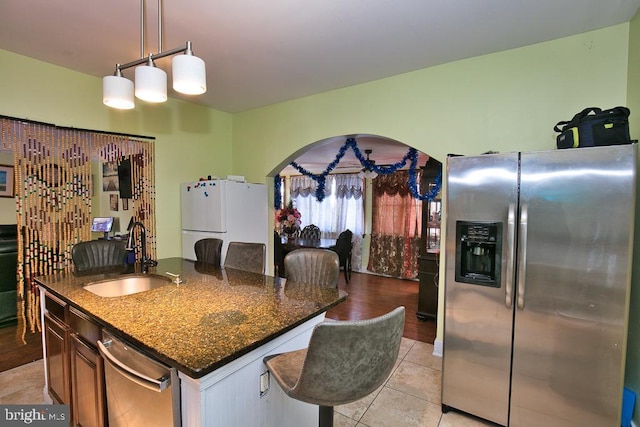 kitchen with arched walkways, light tile patterned floors, appliances with stainless steel finishes, a kitchen island with sink, and a sink