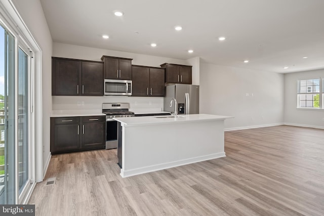 kitchen with an island with sink, appliances with stainless steel finishes, dark brown cabinetry, and light hardwood / wood-style flooring