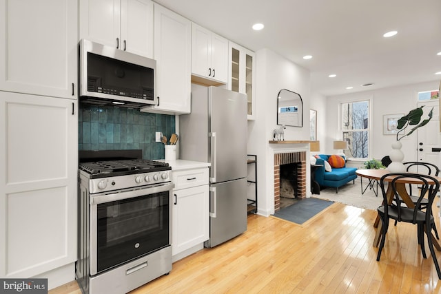 kitchen with a fireplace, stainless steel appliances, light wood-style flooring, decorative backsplash, and white cabinetry