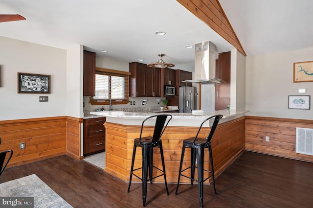 kitchen featuring stainless steel appliances, a kitchen breakfast bar, island range hood, kitchen peninsula, and wood walls