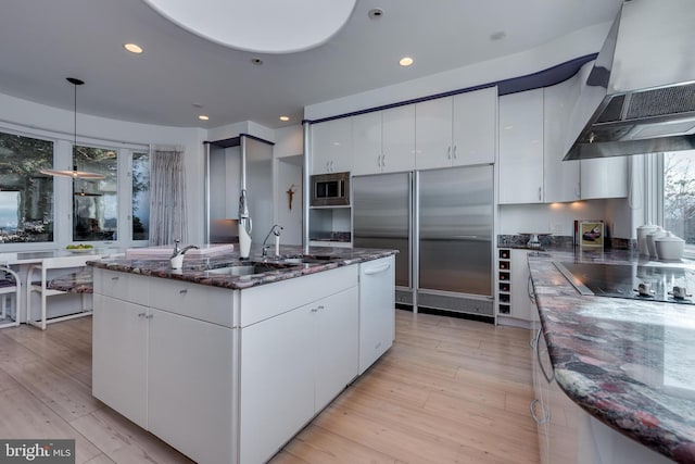 kitchen with white cabinets, dark stone counters, built in appliances, and exhaust hood