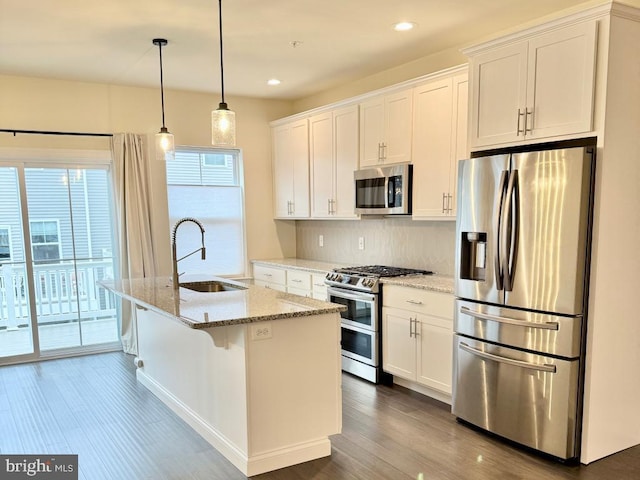 kitchen featuring appliances with stainless steel finishes, pendant lighting, white cabinetry, an island with sink, and light stone counters