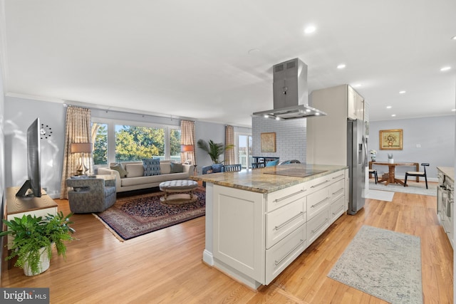 kitchen with open floor plan, white cabinetry, island range hood, light stone countertops, and stainless steel fridge