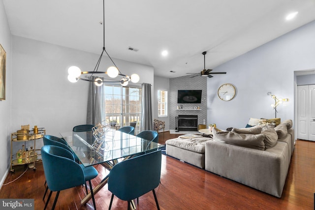 dining room with visible vents, ceiling fan with notable chandelier, wood finished floors, a brick fireplace, and vaulted ceiling