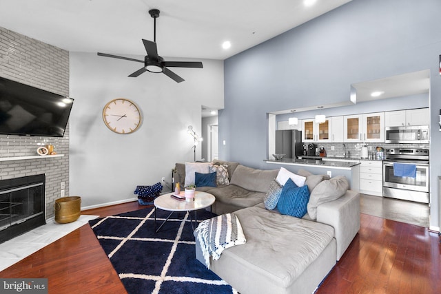 living area with a brick fireplace, baseboards, dark wood finished floors, high vaulted ceiling, and a ceiling fan