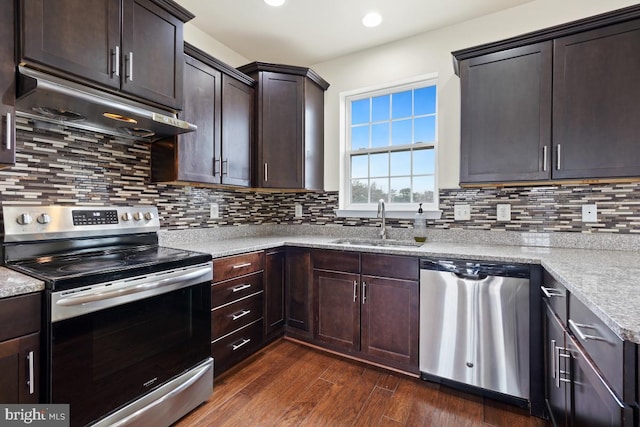 kitchen with under cabinet range hood, stainless steel appliances, a sink, light stone countertops, and dark wood-style floors