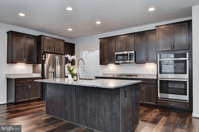 kitchen featuring stainless steel appliances, dark brown cabinetry, light stone countertops, an island with sink, and dark hardwood / wood-style flooring