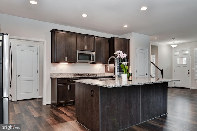 kitchen featuring sink, appliances with stainless steel finishes, a kitchen island with sink, dark brown cabinetry, and light stone countertops