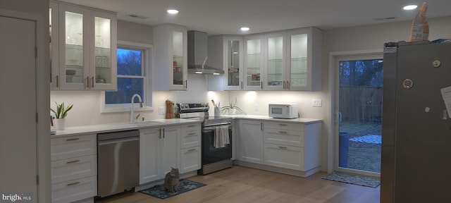 kitchen featuring sink, light wood-type flooring, appliances with stainless steel finishes, wall chimney range hood, and white cabinets