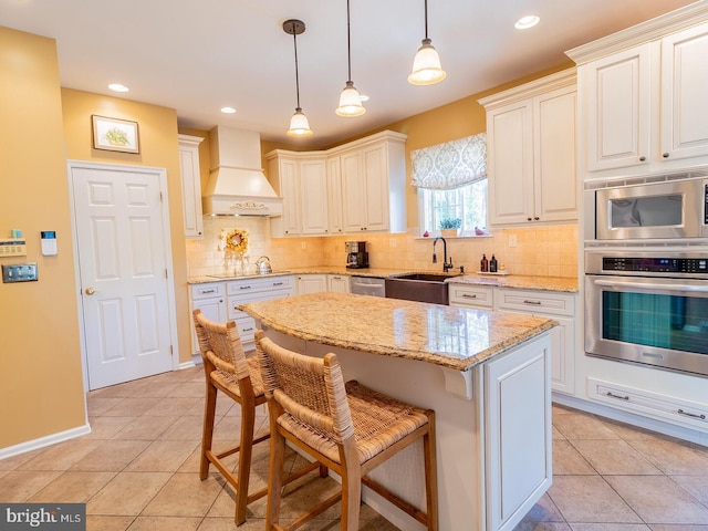 kitchen with sink, stainless steel appliances, light stone counters, custom range hood, and decorative light fixtures