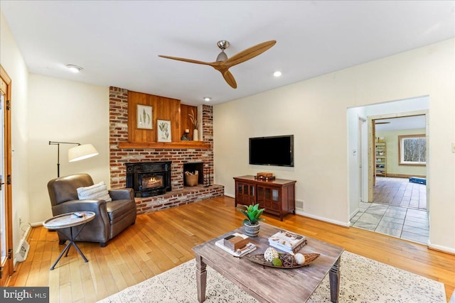 living room featuring ceiling fan, wood-type flooring, baseboards, and a brick fireplace