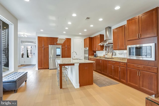 kitchen featuring wall chimney exhaust hood, appliances with stainless steel finishes, a kitchen breakfast bar, an island with sink, and light stone countertops