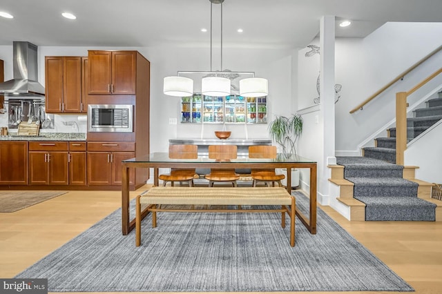 kitchen with hanging light fixtures, wall chimney range hood, stainless steel microwave, and light hardwood / wood-style floors