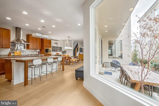 kitchen with pendant lighting, stainless steel microwave, an island with sink, light stone counters, and wall chimney exhaust hood