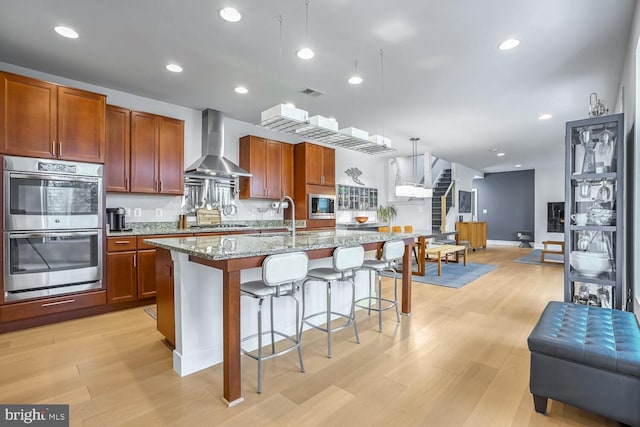 kitchen featuring appliances with stainless steel finishes, decorative light fixtures, an island with sink, light stone counters, and wall chimney range hood