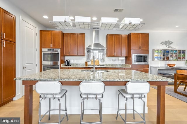 kitchen featuring appliances with stainless steel finishes, decorative light fixtures, a center island with sink, and wall chimney range hood