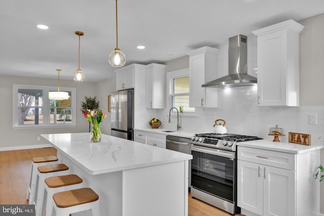 kitchen featuring wall chimney exhaust hood, a breakfast bar area, decorative light fixtures, appliances with stainless steel finishes, and white cabinets