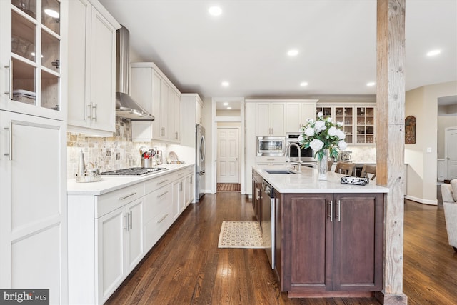 kitchen with tasteful backsplash, dark wood finished floors, stainless steel appliances, wall chimney range hood, and glass insert cabinets