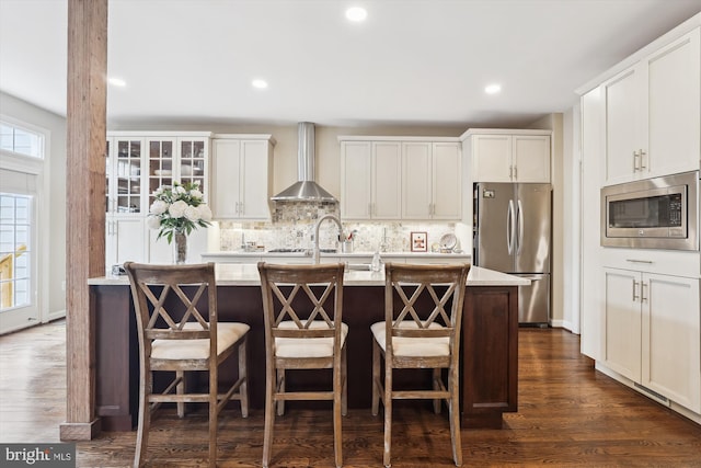 kitchen featuring dark wood-type flooring, a kitchen island with sink, appliances with stainless steel finishes, wall chimney range hood, and decorative backsplash