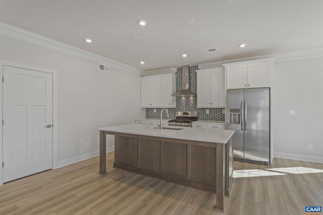 kitchen featuring wall chimney exhaust hood, sink, appliances with stainless steel finishes, an island with sink, and white cabinets