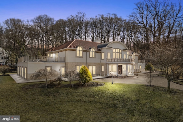 back house at dusk featuring a patio, a balcony, and a lawn