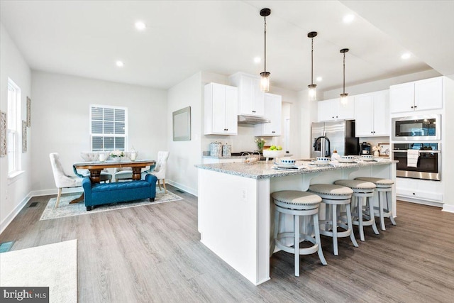 kitchen with white cabinets, appliances with stainless steel finishes, light stone counters, hanging light fixtures, and a kitchen island with sink