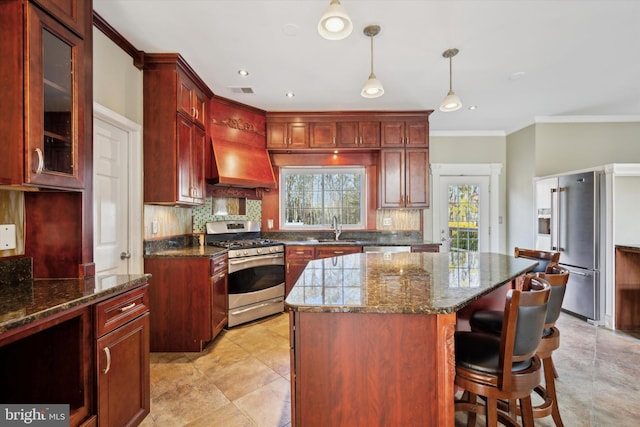 kitchen with a kitchen island, appliances with stainless steel finishes, dark stone counters, hanging light fixtures, and custom range hood