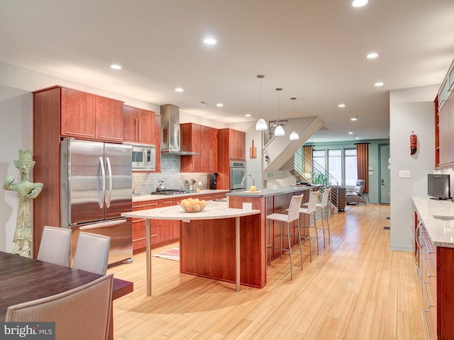 kitchen featuring light wood-type flooring, a kitchen breakfast bar, pendant lighting, stainless steel appliances, and wall chimney range hood