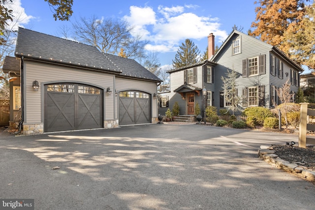 view of front of house with an attached garage, driveway, stone siding, roof with shingles, and a chimney