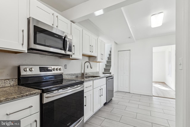 kitchen with appliances with stainless steel finishes, white cabinets, a sink, dark stone counters, and baseboards