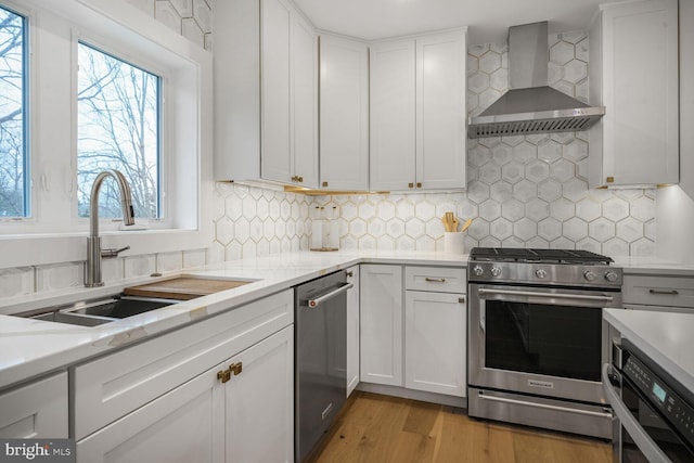 kitchen with stainless steel appliances, light countertops, wall chimney range hood, white cabinetry, and a sink